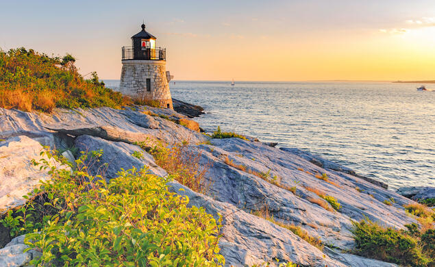 A senice view of a rocky ledge leading out to a Castle Hill Lighthouse and a view of the water at sunset.