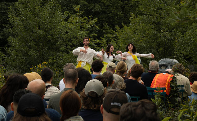 Three actors perform on a stage outside before a small audience.
