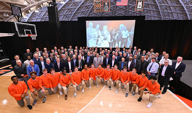 A large group of alumni stand behind the current men's basketball team, who are on one knee. In the background is a large screen showing photos of Pete Carril.