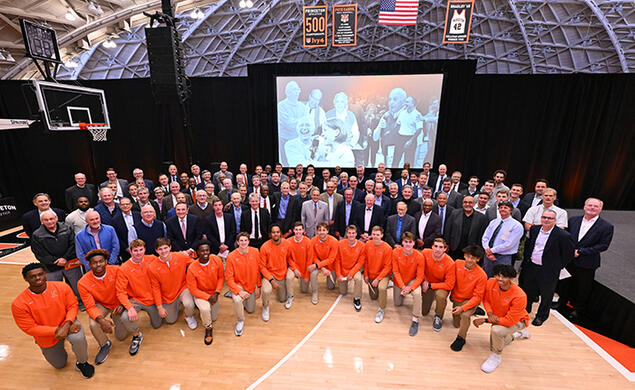 A large group of alumni stand behind the current men's basketball team, who are on one knee. In the background is a large screen showing photos of Pete Carril.