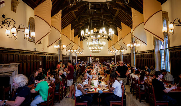 Students sit at long tables arranged in three rows in a dining hall with a high wood ceiling.