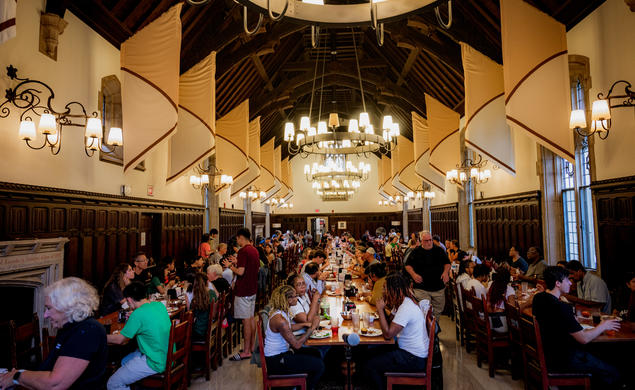 Students sit at long tables arranged in three rows in a dining hall with a high wood ceiling.