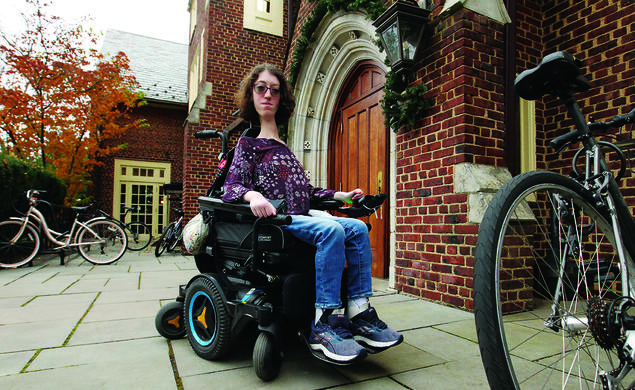 This is a photo of Naomi Hess ’22 in her motorized wheelchair in front of a Princeton campus building. Bicycles are in the foreground and background.