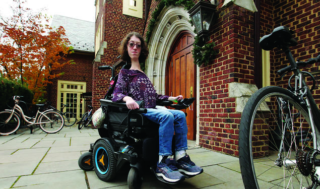 This is a photo of Naomi Hess ’22 in her motorized wheelchair in front of a Princeton campus building. Bicycles are in the foreground and background.