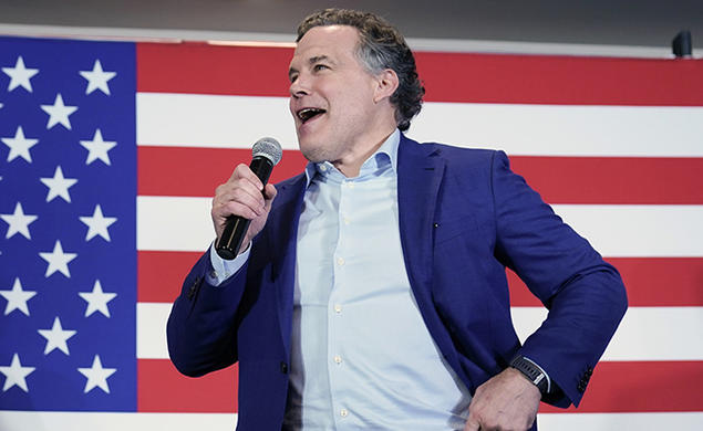 This is a photo of David McCormick speaking into a microphone, with a large American flag in the background.