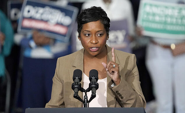 Andrea Campbell speaks at a podium; people hold campaign signs in the background.
