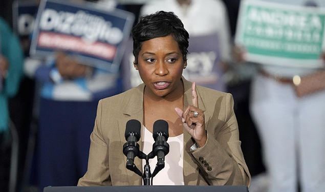 Andrea Campbell speaks at a podium; people hold campaign signs in the background.