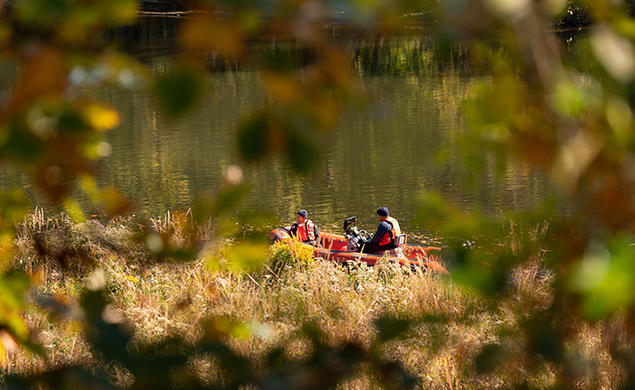 Seen through an opening in the trees, two people in orange vests motor on Lake Carnegie in an orange boat.