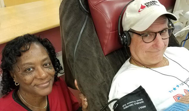 Wearing a "Virginia Blood" baseball cap, Doug Schutte ’76 sits in a chair wearing headphones and a blood pressure monitor as a Red Cross worker poses next to him.