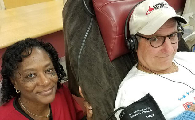 Wearing a "Virginia Blood" baseball cap, Doug Schutte ’76 sits in a chair wearing headphones and a blood pressure monitor as a Red Cross worker poses next to him.