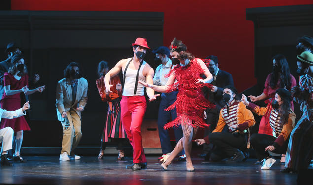 This is a photo of students on stage during a Triangle Club rehearsal. A boy and a girl dance in front while others snap their fingers while watching in back and on the sides. All are wearing black KN95 masks.