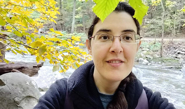 Elizabeth Tsurkov takes a selfie under a tree with a small river behind her.