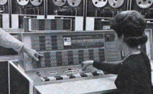 A woman sits at the controls of the room-sized IBM 7090 at Princeton’s Computer Center, manually entering instructions. You can see the data storage tapes at the top of the screen.