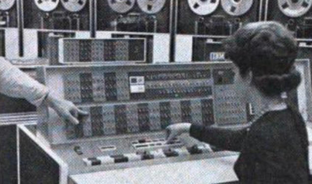 A woman sits at the controls of the room-sized IBM 7090 at Princeton’s Computer Center, manually entering instructions. You can see the data storage tapes at the top of the screen.