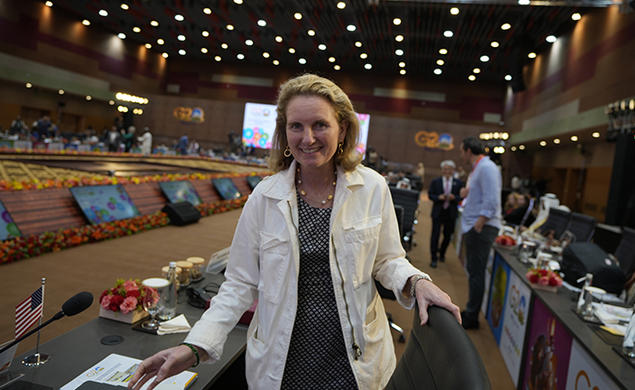 Isobel Coleman ’87 faces the camera and smiles in a room where each seat has a small screen. Blurry in the background are lit-up signs reading G20.