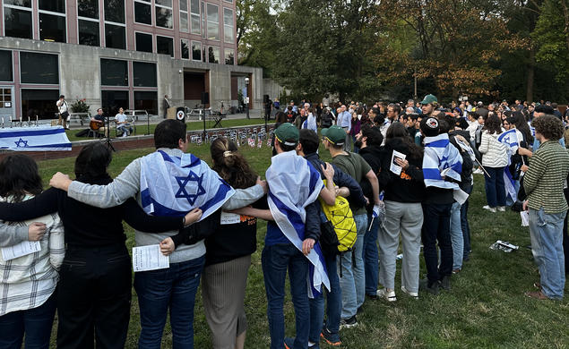People stand with their arms around each other's shoulders outside a campus building, listening to two students play music. A few have blue-and-white Israel flags wrapped around their shoulders.