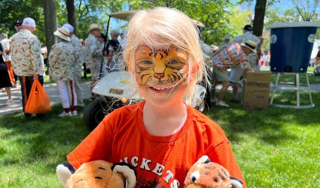 A six-year-old girl with tiger faceprint, an orange shirt, and a plush tiger under each arm.