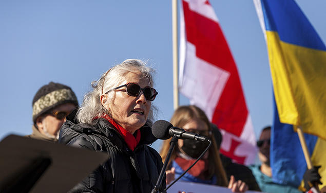 Former U.S. ambassador to Ukraine Marie Yovanovitch ’80 speaks into a microphone during a solidarity rally for Ukraine on Feb. 19, outdoors, wearing sunglasses, with blue sky and the Ukrainian flag in the background.