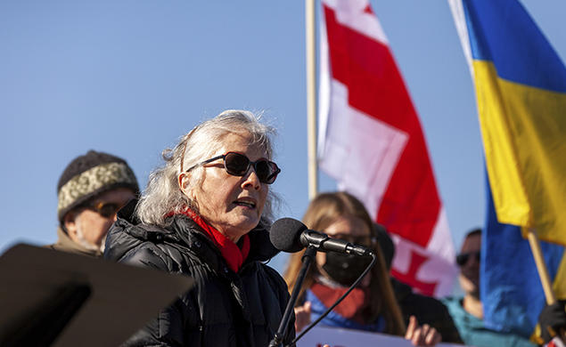 Former U.S. ambassador to Ukraine Marie Yovanovitch ’80 speaks into a microphone during a solidarity rally for Ukraine on Feb. 19, outdoors, wearing sunglasses, with blue sky and the Ukrainian flag in the background.
