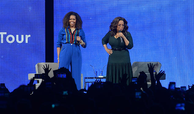 Michelle Obama and Oprah address an audience from a stage.