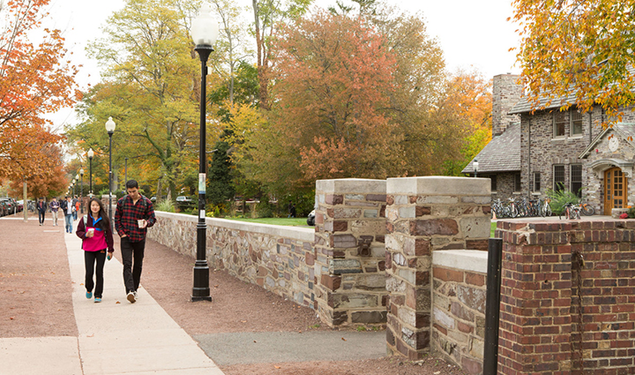 Two students holding coffee cups walk on a sidewalk next to a stone wall. A stone eating club can be seen to the right behind the wall.