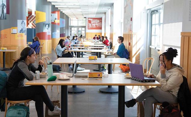 This is a photo of students working on laptops and eating in the Frist Campus Center, distanced by sitting longways across from each other, two to a table.
