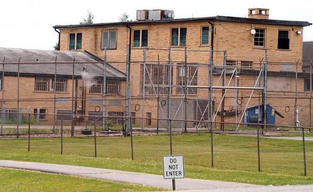 This is a photo of the Edna Mahan Correctional Facility, made of yellow brick with tall chain-link fences in front and a small sign reading "DO NOT ENTER."