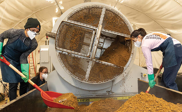 Students shovel compost that looks like dirt next to a cylindrical machine.