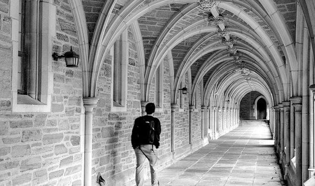 A black-and-white photo of a student walking down an arched stone hallway alone.