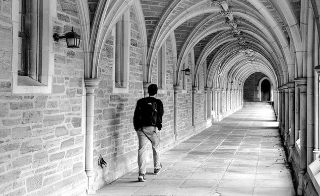 A black-and-white photo of a student walking down an arched stone hallway alone.