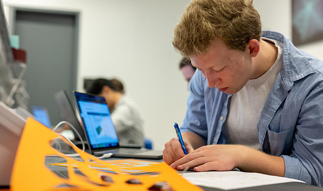 A student writes with a pen; a yellow piece of paper with shapes punched out is in the foreground.