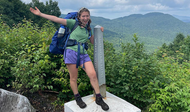 Wearing her backpack and hiking boots, Ruby Platt flings out an arm and smiles as she stands in front of the Green Mountains, holding onto an obelisk-shaped marker that reads "Treaty of Washington."