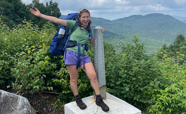 Wearing her backpack and hiking boots, Ruby Platt flings out an arm and smiles as she stands in front of the Green Mountains, holding onto an obelisk-shaped marker that reads "Treaty of Washington."