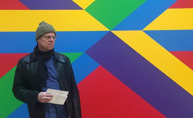 This is a photo of Joe Scanlan holding papers and standing in front of a brightly-painted wall.