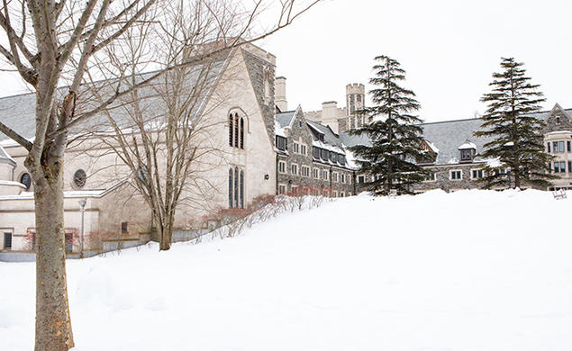 A view of part of Princeton’s Gothic stone campus, viewed up a snowy hill, with two large pine trees at the top.