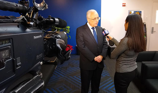 This is a photo of Alain Sanders being interviewed by a news reporter holding a microphone. A video camera filming them is in the foreground..