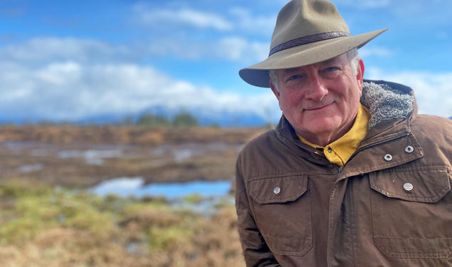 This is a photo of Bill Urschel ’78 with blue sky and an Alaskan summer landscape in the background.