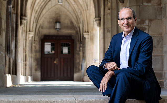 This is a photo of David Nirenberg sitting on the steps of a building with Gothic arches in the background.