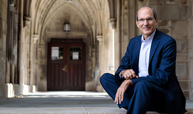 This is a photo of David Nirenberg sitting on the steps of a building with Gothic arches in the background.