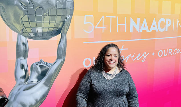 Janice Johnston ’92 stands in front of a large pink-and-yellow sign reading "54th NAACP." Part of the text is cut off. There's also a photo of a silver statue of Atlas holding up the globe.