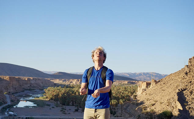Wearing a blue shirt and a backpack, Ryan Ozminkowski looks up as he juggles rocks. A canyon is in the background.