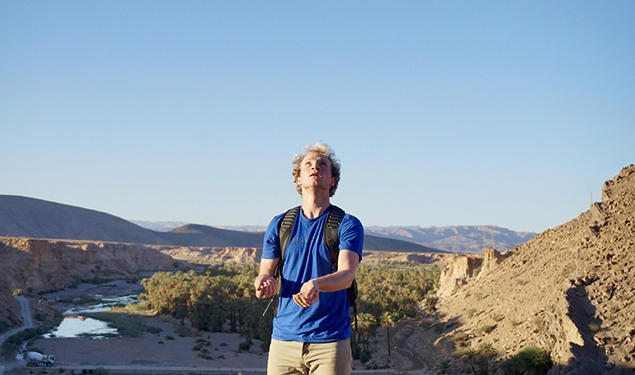Wearing a blue shirt and a backpack, Ryan Ozminkowski looks up as he juggles rocks. A canyon is in the background.