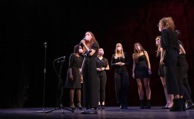 This is a photo of the Tigerlilies singing on stage; eight women are wearing black clothes and pink face masks while they sing.