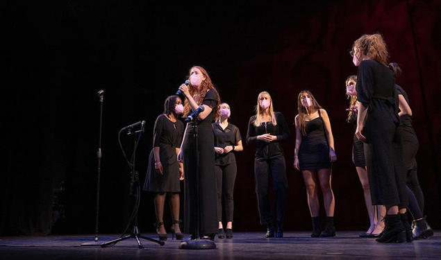 This is a photo of the Tigerlilies singing on stage; eight women are wearing black clothes and pink face masks while they sing.