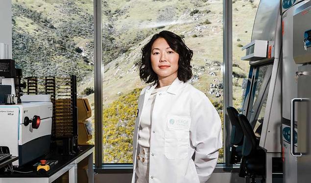 This is a photo of Alice Zhang ’10 inside the Verge Genomics laboratory in South San Francisco, wearing a lab coat and surrounded by machines, with a view of scrubby vegetation through the window behind her.