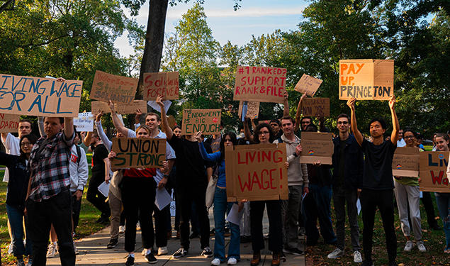 A crowd standing outside holds signs reading "Living Wage!" and "Pay Up Princeton" and #1 Ranked? Support Rank & File" and "Union Strong."