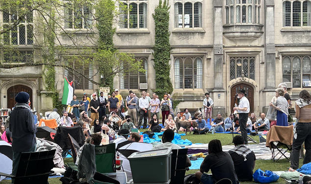 A man stands at a microphone surrounded by protesters; a Gothic building is in the background.