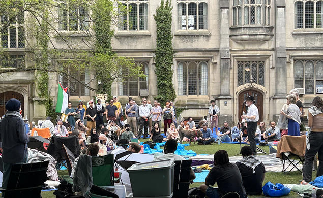 A man stands at a microphone surrounded by protesters; a Gothic building is in the background.