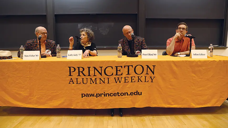 Four people sit at a long tabble draped with an orange cloth. 