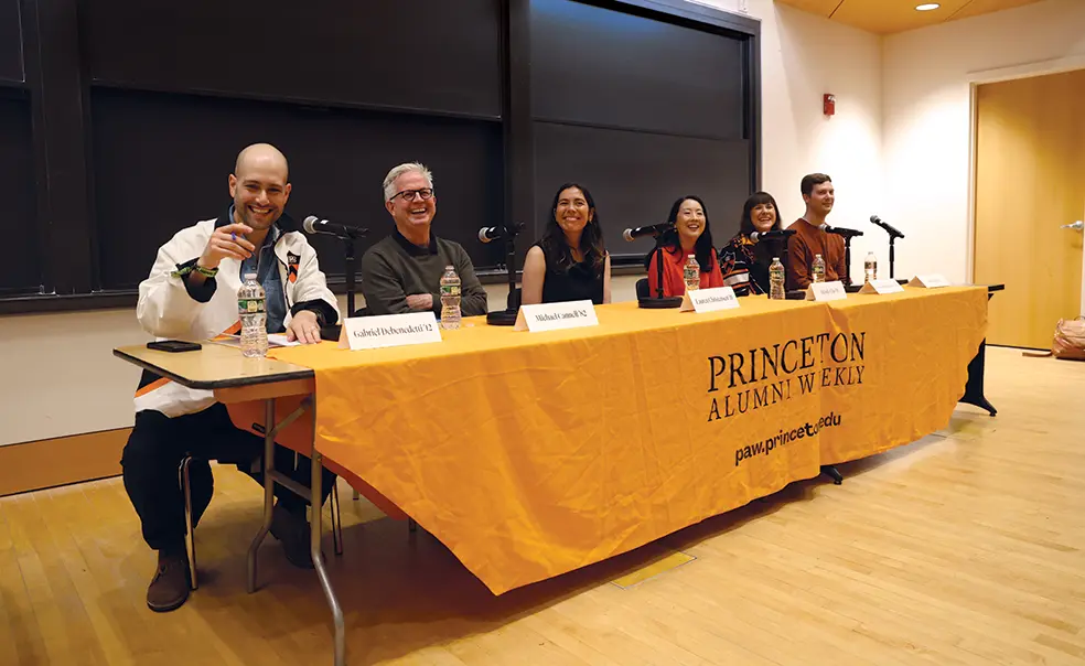 Six people sit at a table in the front of a classroom; the table has an orange tablecloth that reads Princeton Alumni Weekly.
